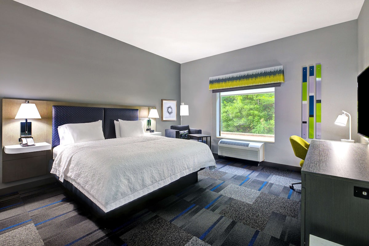 A modern hotel room with a large bed, bedside tables with lamps, a desk, and a chair. There is a window with a view of greenery outside.