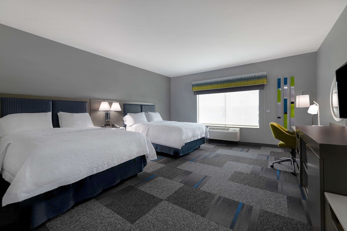 A hotel room with two beds, a desk, a chair, a window, and a radiator.