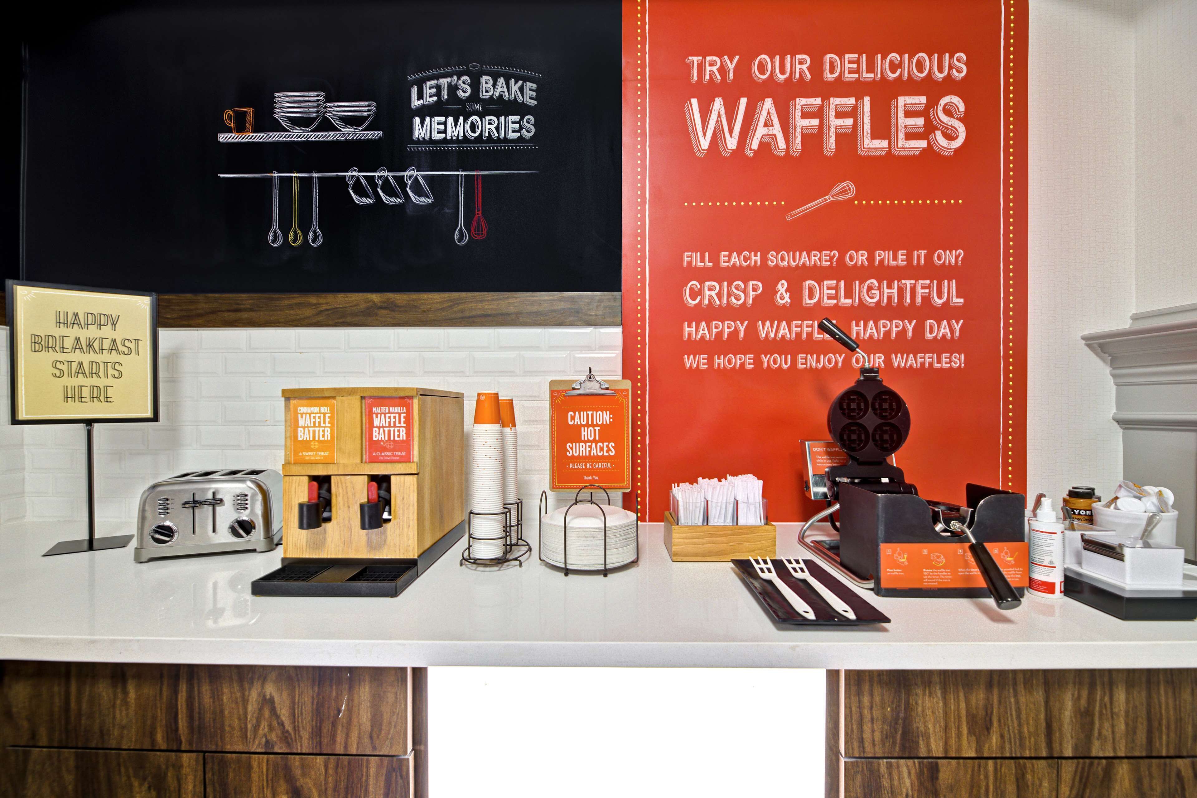 A breakfast setup with waffle batter dispensers, a waffle maker, and a sign indicating breakfast starts here.