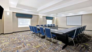 A conference room with a long table, blue chairs, a projector screen, and large windows.