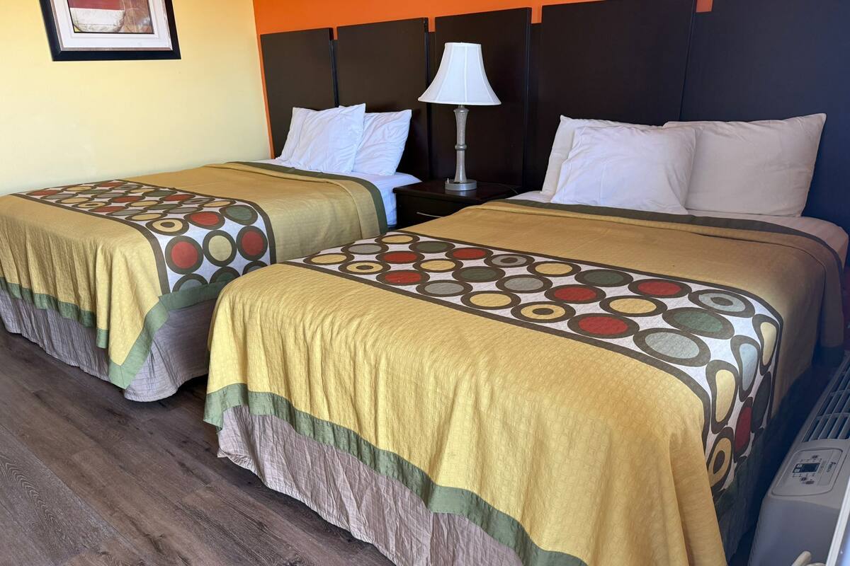 Dos camas dobles con colchas estampadas, una mesita de noche con lámpara y un calentador en el suelo.