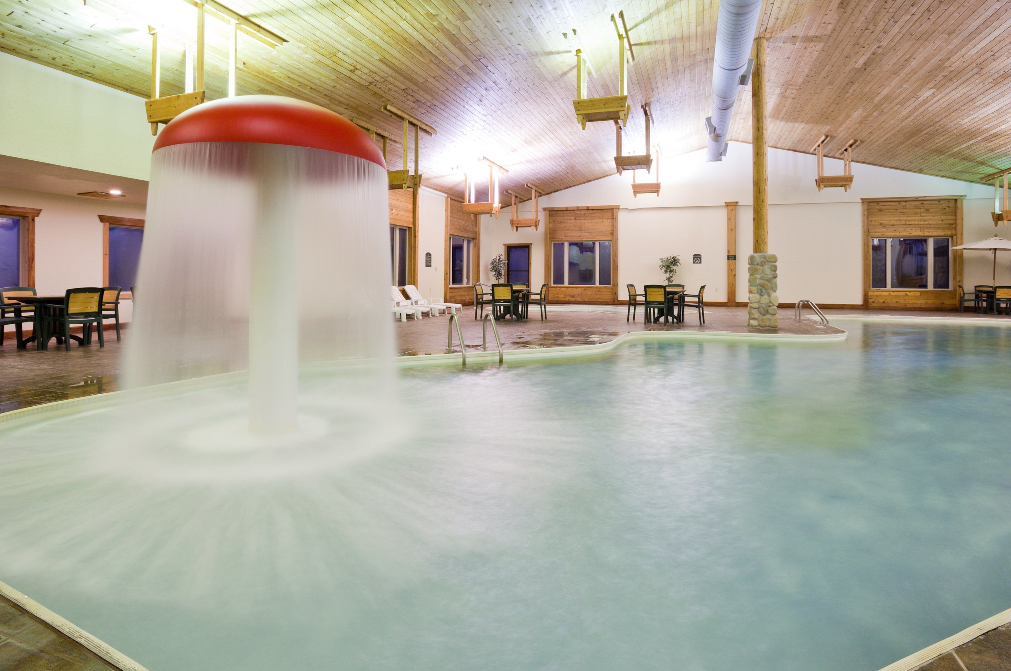Una amplia piscina interior con numerosas tumbonas y mesas, vigas de madera en el techo y un pilar de piedra.