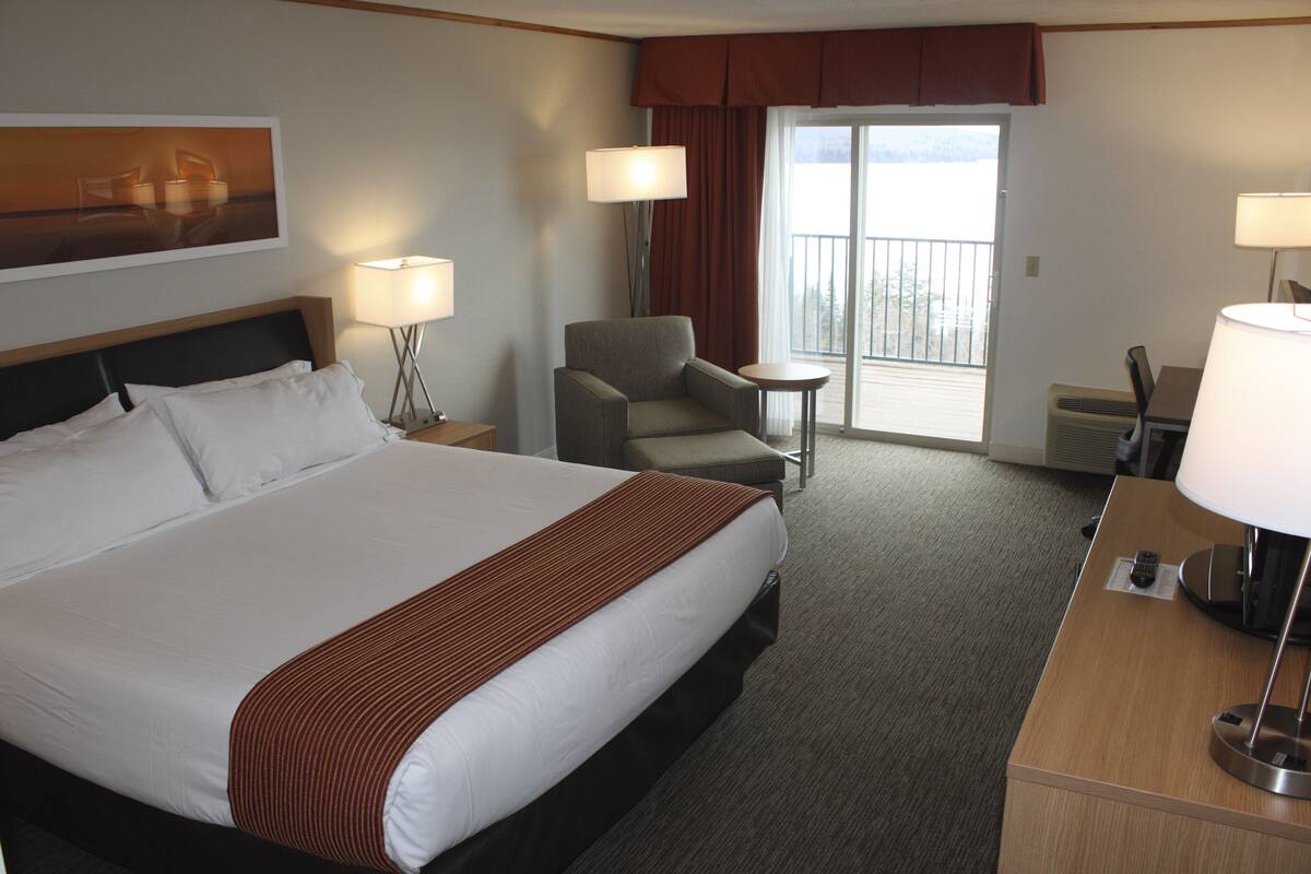 A hotel room with a large bed, a chair, a small table, and a desk with a lamp.