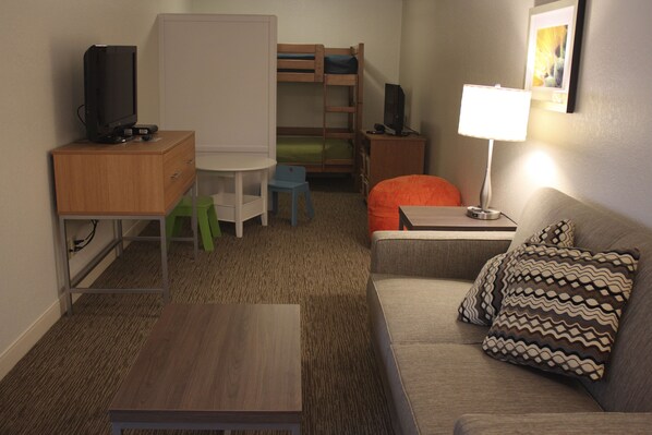Habitación de hotel compacta con literas, un escritorio con computadora, una mesita pequeña, un sofá con cojines estampados y una lámpara de noche.
