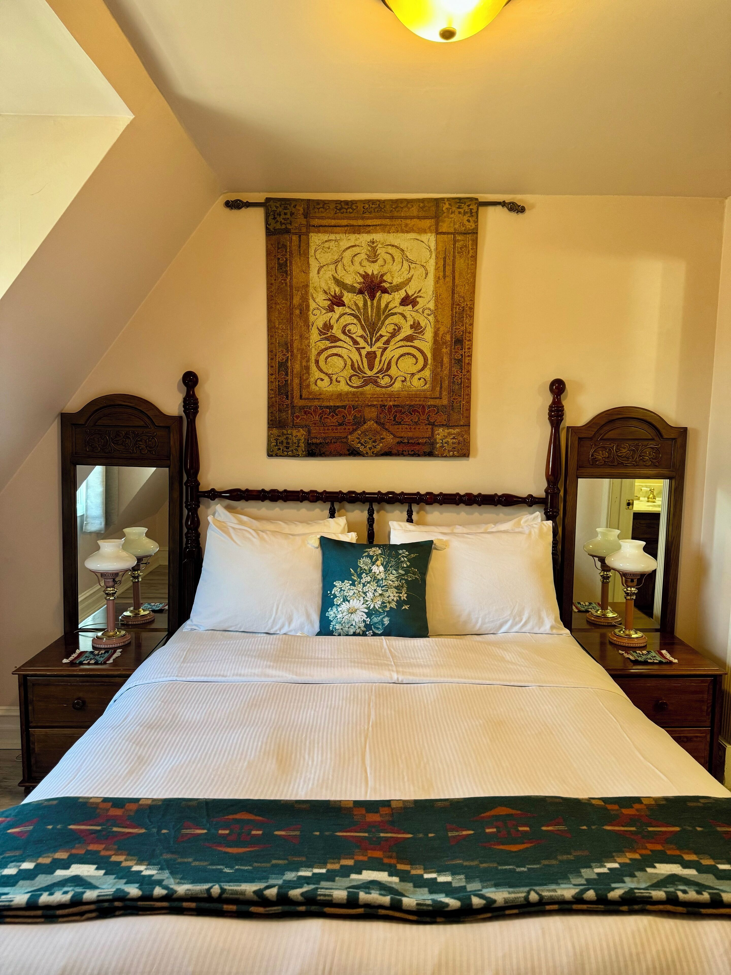 A neatly made bed with white linens, a green floral pillow, and a patterned blanket. There are two bedside tables with lamps and a large mirror above the bed.