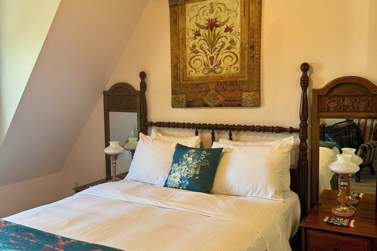 A neatly made bed with a patterned throw, a wooden nightstand with a lamp, and a large tapestry on the wall.