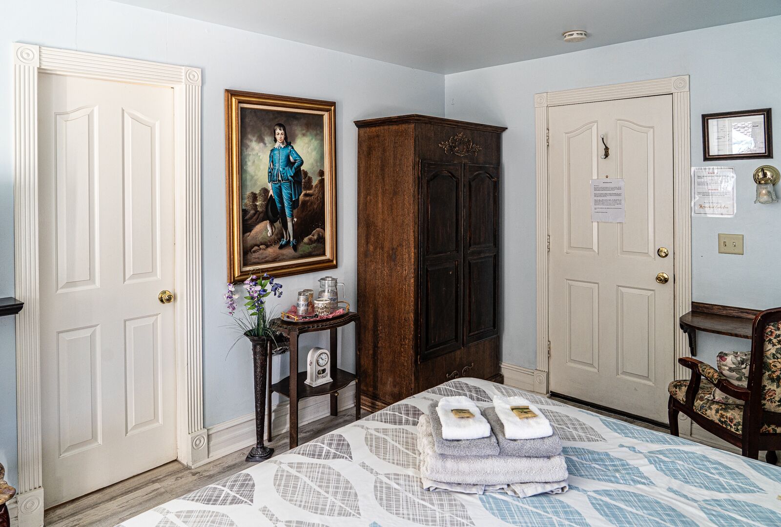 A bedroom with a bed, a dresser, a nightstand, a painting, and a door with a notice.