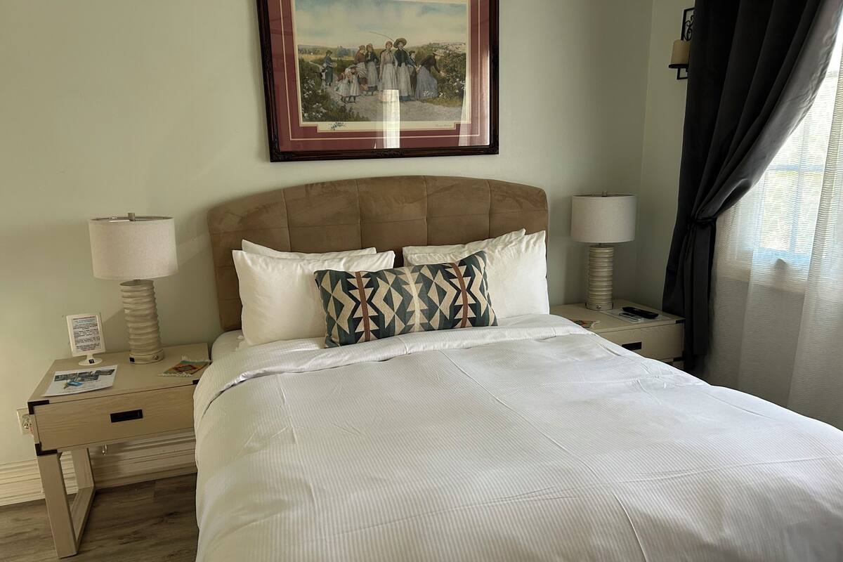 A neatly made bed with white linens, a headboard, and two decorative pillows. There is a framed picture on the wall above the bed, a bedside lamp, and a nightstand with a lamp and a small table.