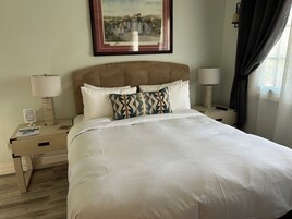 Un letto ben rifatto con lenzuola bianche, una testiera e due cuscini decorativi. Sopra il letto, una cornice con un quadro, una lampada da comodino e un comodino con una lampada e un piccolo tavolino.