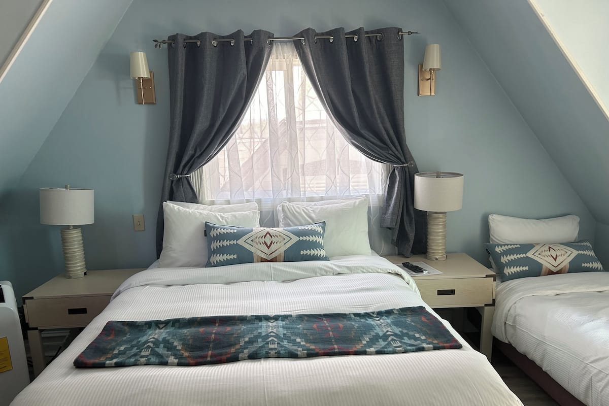 A bedroom with a sloped ceiling, two bedside tables with lamps, a bed with white bedding and patterned pillows, and a window with curtains.