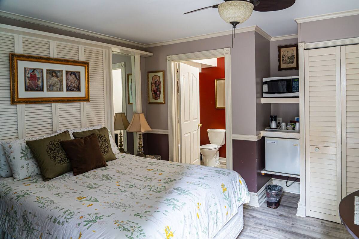 A bedroom with a bed, bedside lamps, a ceiling fan, a microwave, and a small refrigerator.
