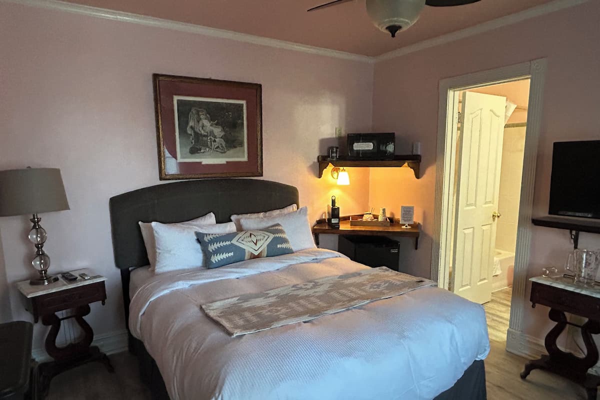 A neatly arranged bedroom with a bed, bedside tables, a nightstand, a framed picture, a shelf with items, and a television.