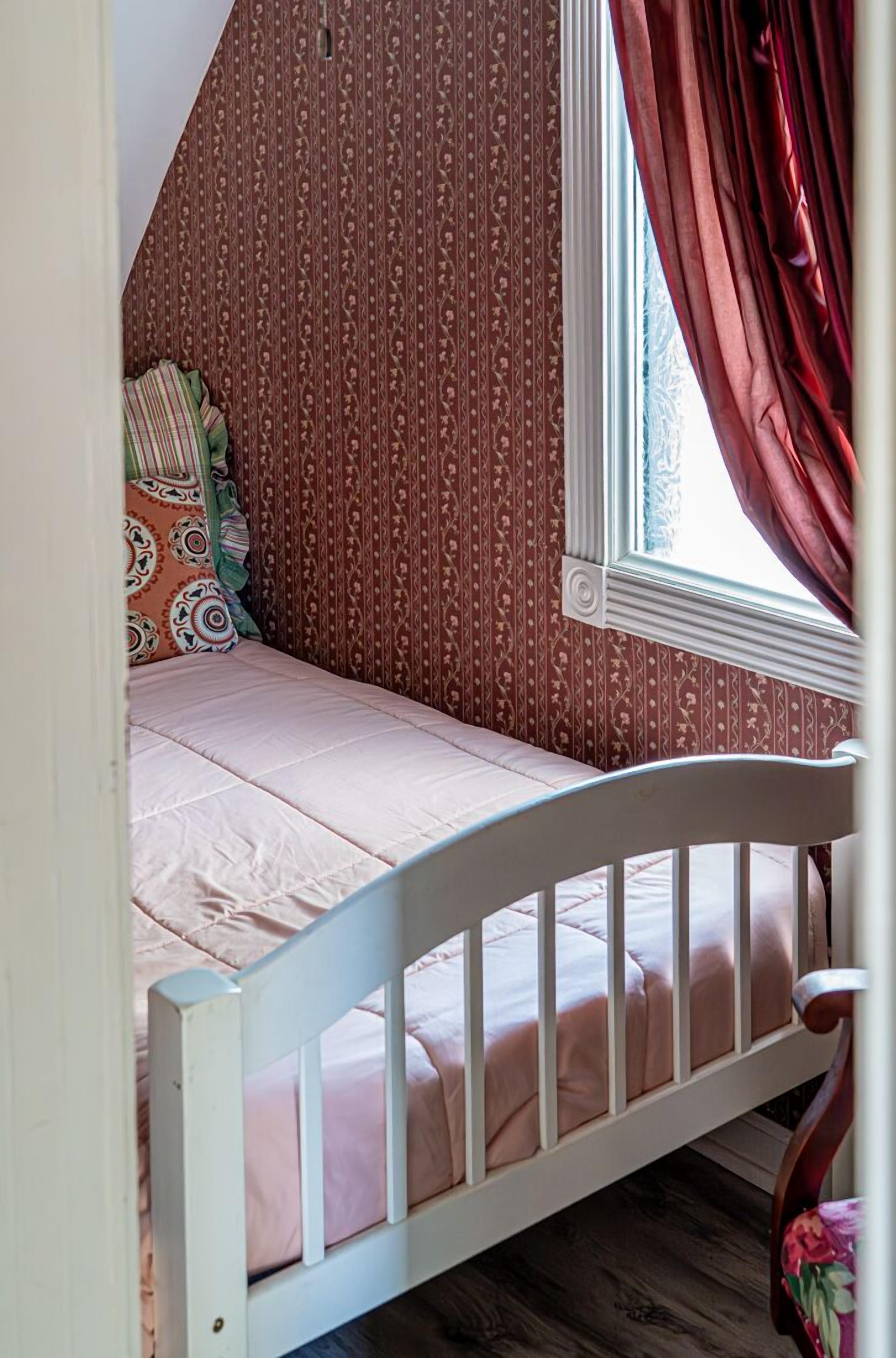 A small, cozy room with a single bed, patterned wallpaper, and a window with curtains.
