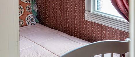 Une petite pièce agréable, dotée d’un lit simple, d’un papier peint à motifs et d’une fenêtre avec des rideaux.