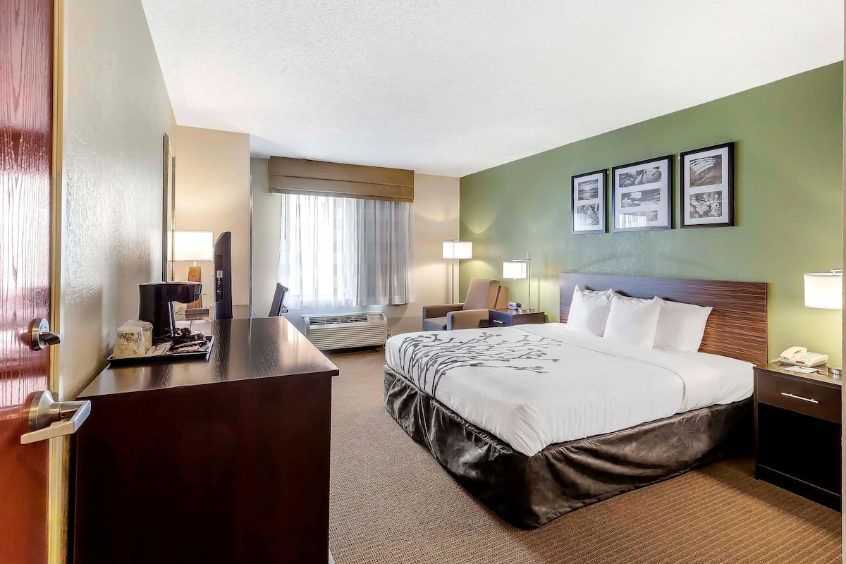 A hotel room with a large bed, a desk with a telephone, a chair, a window with curtains, and a wall with framed pictures.