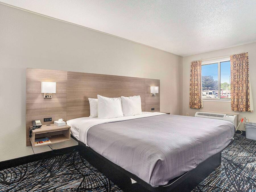 A hotel room with a large bed, bedside lamps, a small table with a phone and tissues, and a window with curtains.
