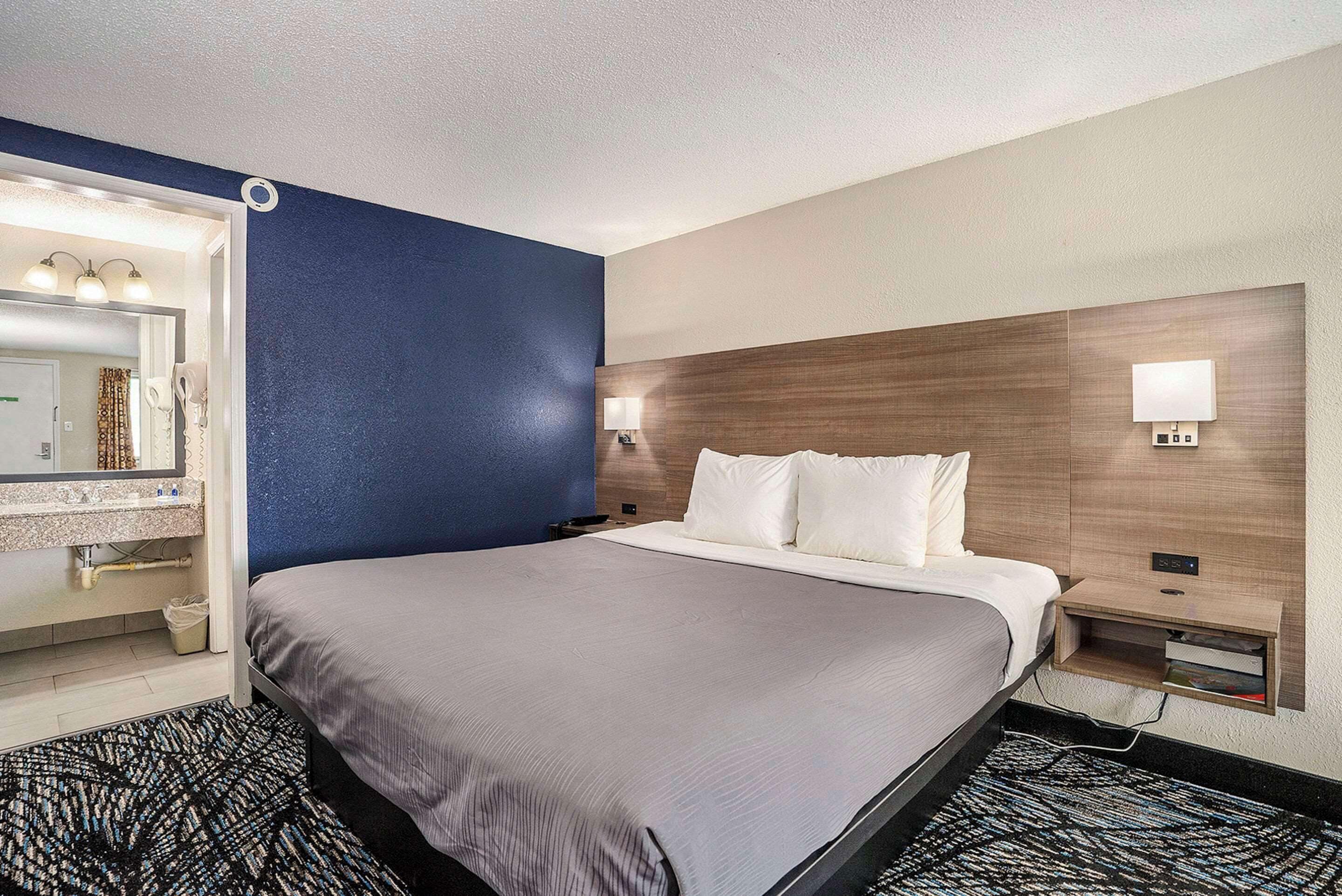 A hotel room with a large bed, a bedside table with a lamp, a mirror, and a wall-mounted shelf.
