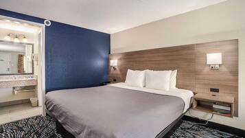 A hotel room with a large bed, a bedside table with a lamp, a mirror, and a wall-mounted shelf.
