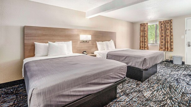 Una habitación de hotel con dos camas, cabeceros, mesitas de noche, una lámpara y una ventana con cortinas.