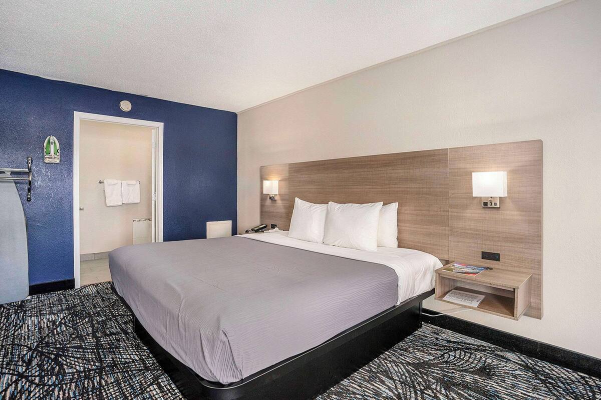 Quarto de hotel com uma cama grande, uma mesa de cabeceira com abajur, uma televisão na parede e uma porta que dá para um banheiro.