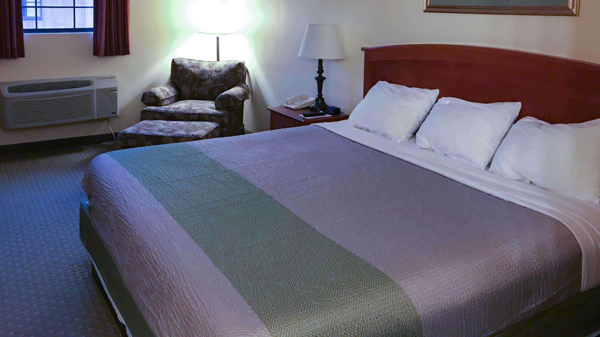 A hotel room with a large bed, a chair, a nightstand with a lamp, and an air conditioning unit.
