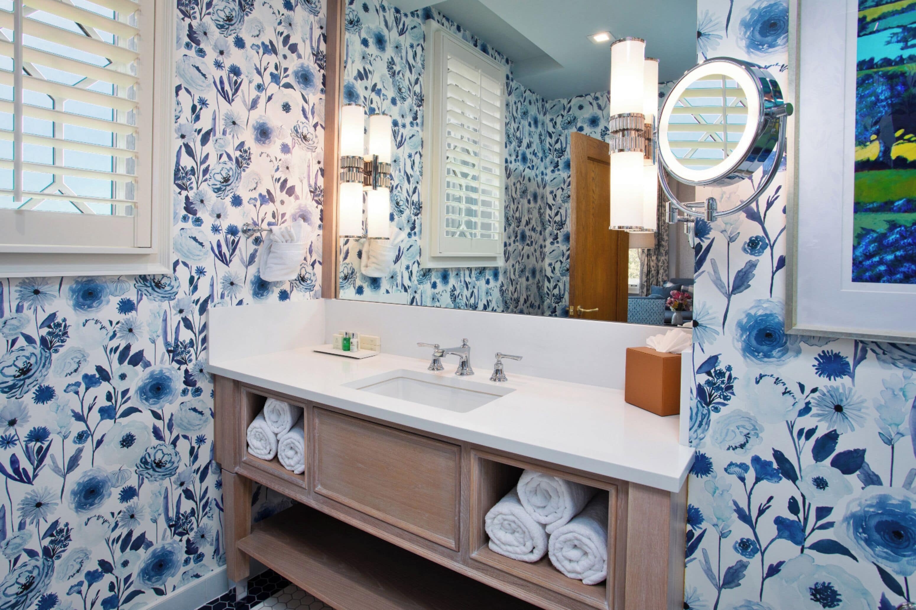 Un baño con papel tapiz floral, un lavamanos blanco, un espejo con iluminación y un toallero.