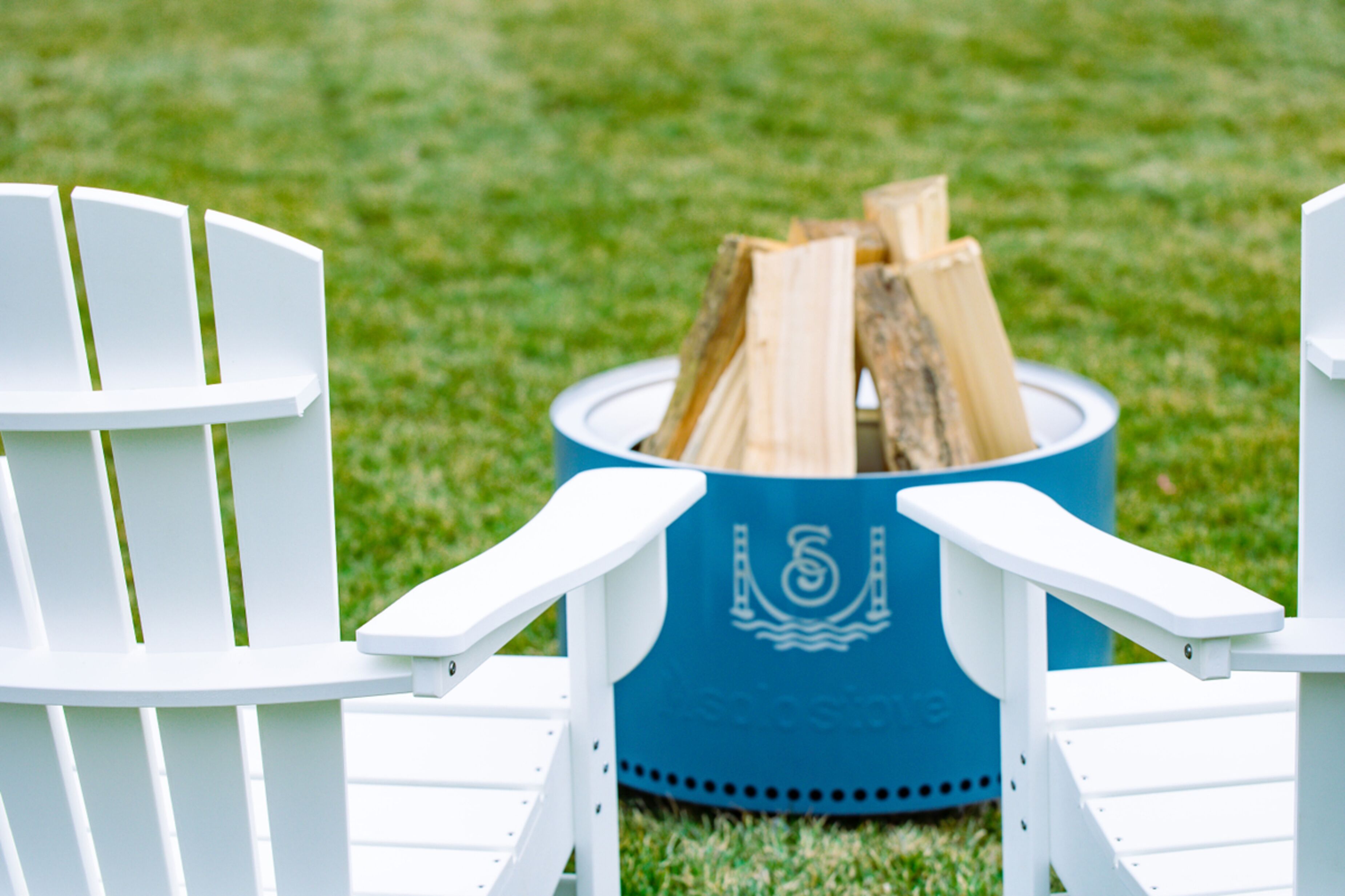 Two white Adirondack chairs facing a blue Adirondack fire pit with logs.