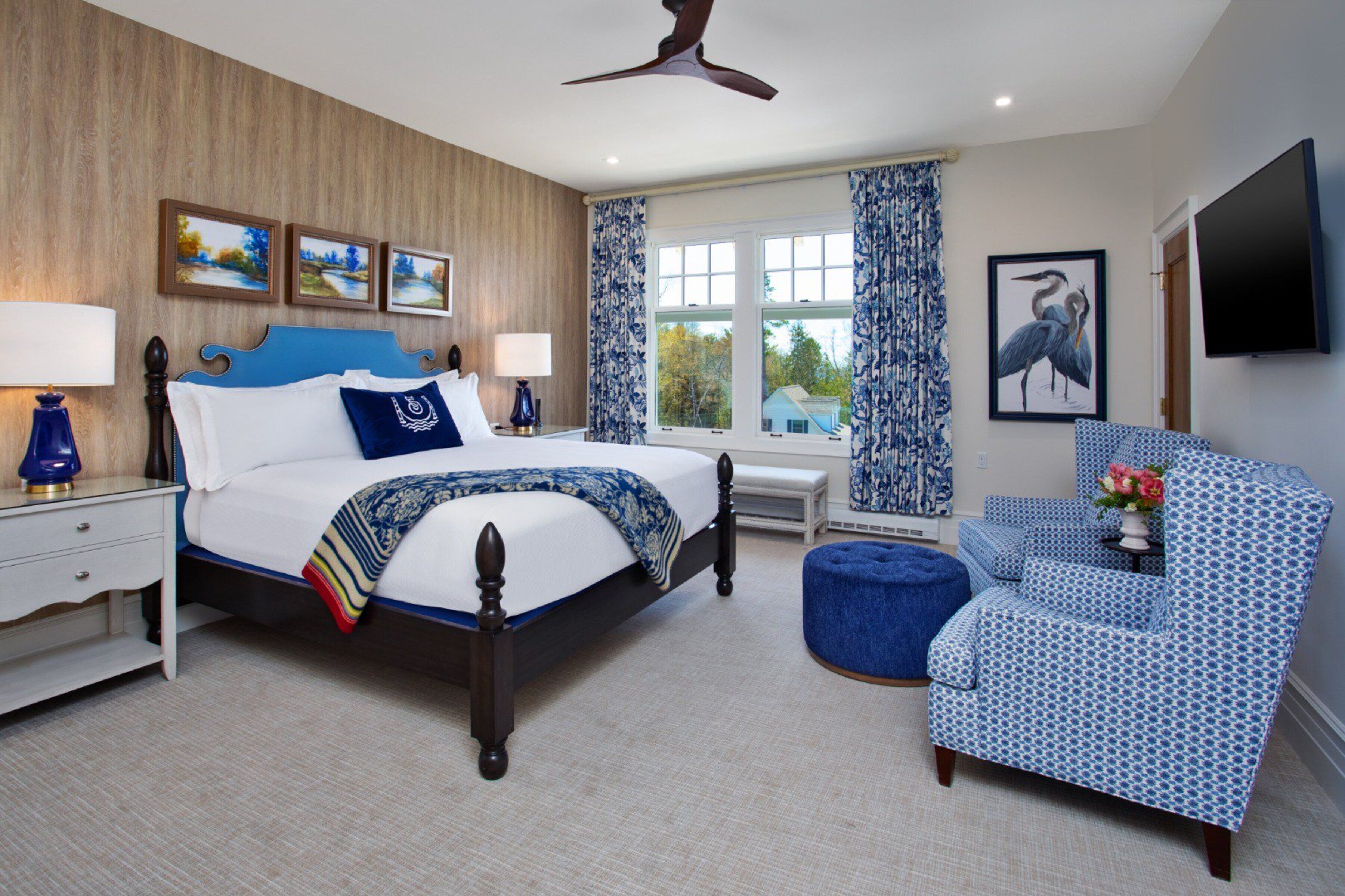 A bedroom with a large bed, a chair, a nightstand, a window with curtains, and a flat-screen TV mounted on the wall.