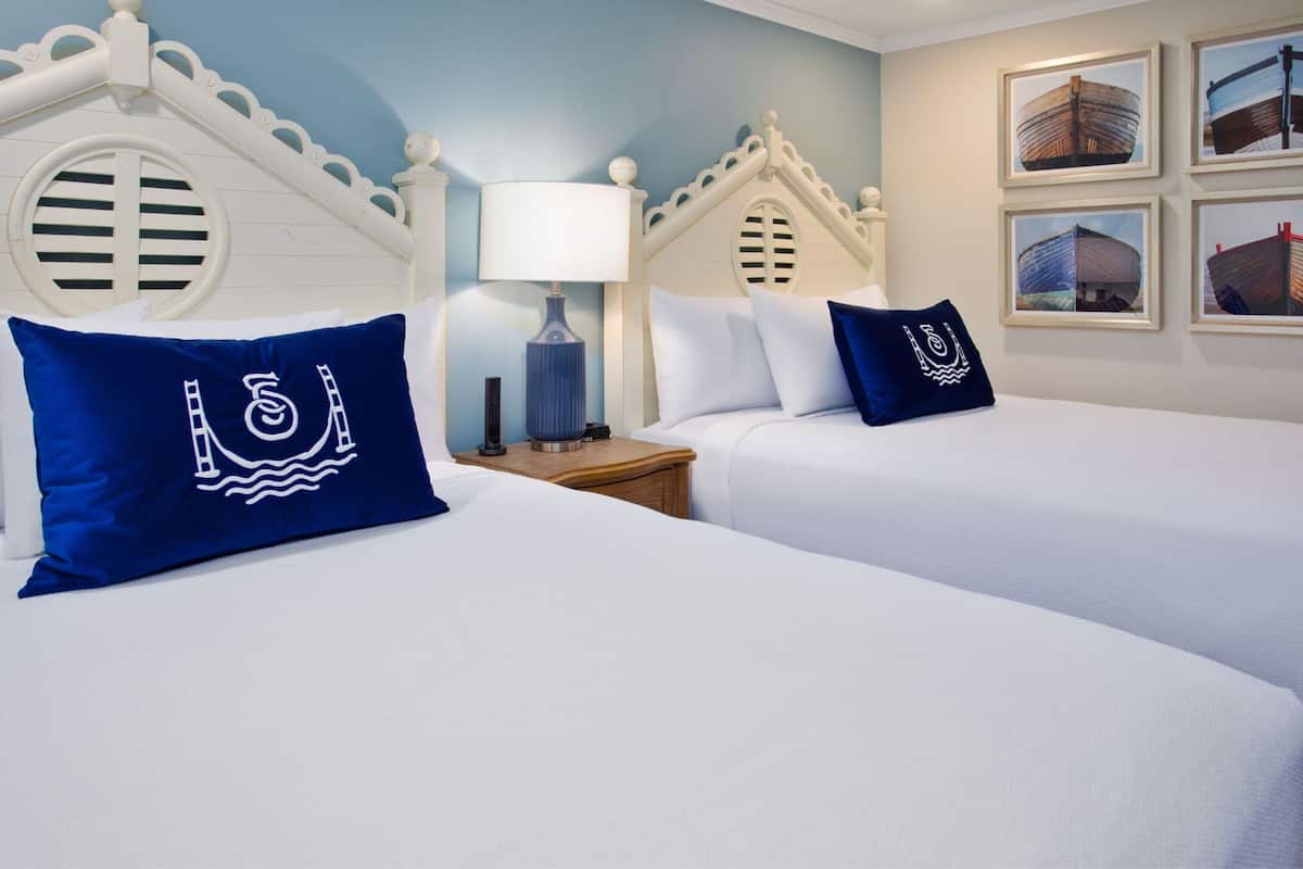 Une chambre d’hôtel avec deux lits, chacun agrémenté de draps blancs et d’oreillers bleus ornés d’un motif nautique. On trouve une table de chevet avec une lampe et une petite table entre les deux lits. Les murs sont peints dans une couleur bleu clair, et des tableaux représentant des bateaux sont accrochés au mur.