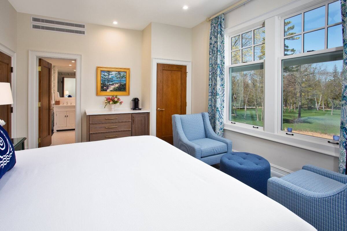 Une chambre à coucher avec un grand lit, une commode en bois, une chaise et une fenêtre donnant sur les arbres.
