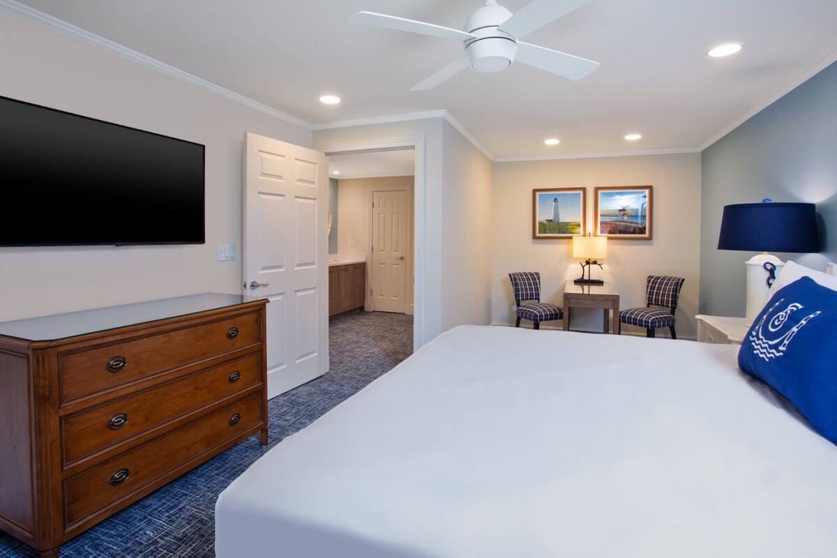 Une chambre d’hôtel avec un grand lit, une commode, un téléviseur à écran plat, deux chaises, une petite table et un ventilateur de plafond.