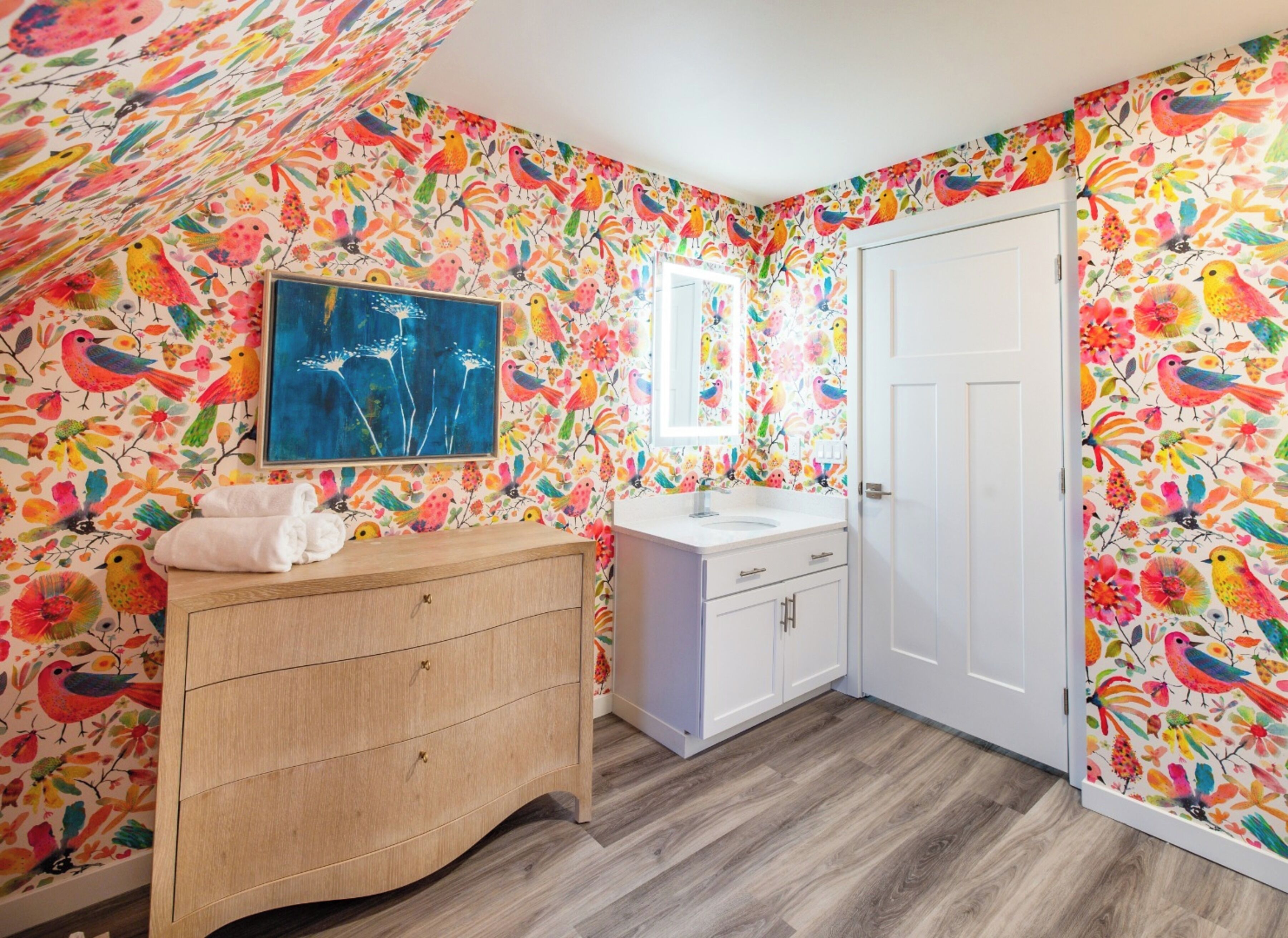 A bathroom with vibrant bird and flower wallpaper, a wooden vanity, a sink, and a door.