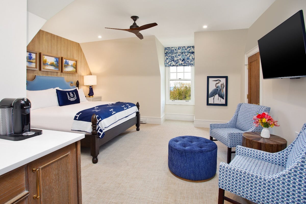 Une chambre d’hôtel comprenant un lit, un bureau, une chaise, un pouf, un ventilateur de plafond, une télévision et des œuvres d’art accrochées aux murs.