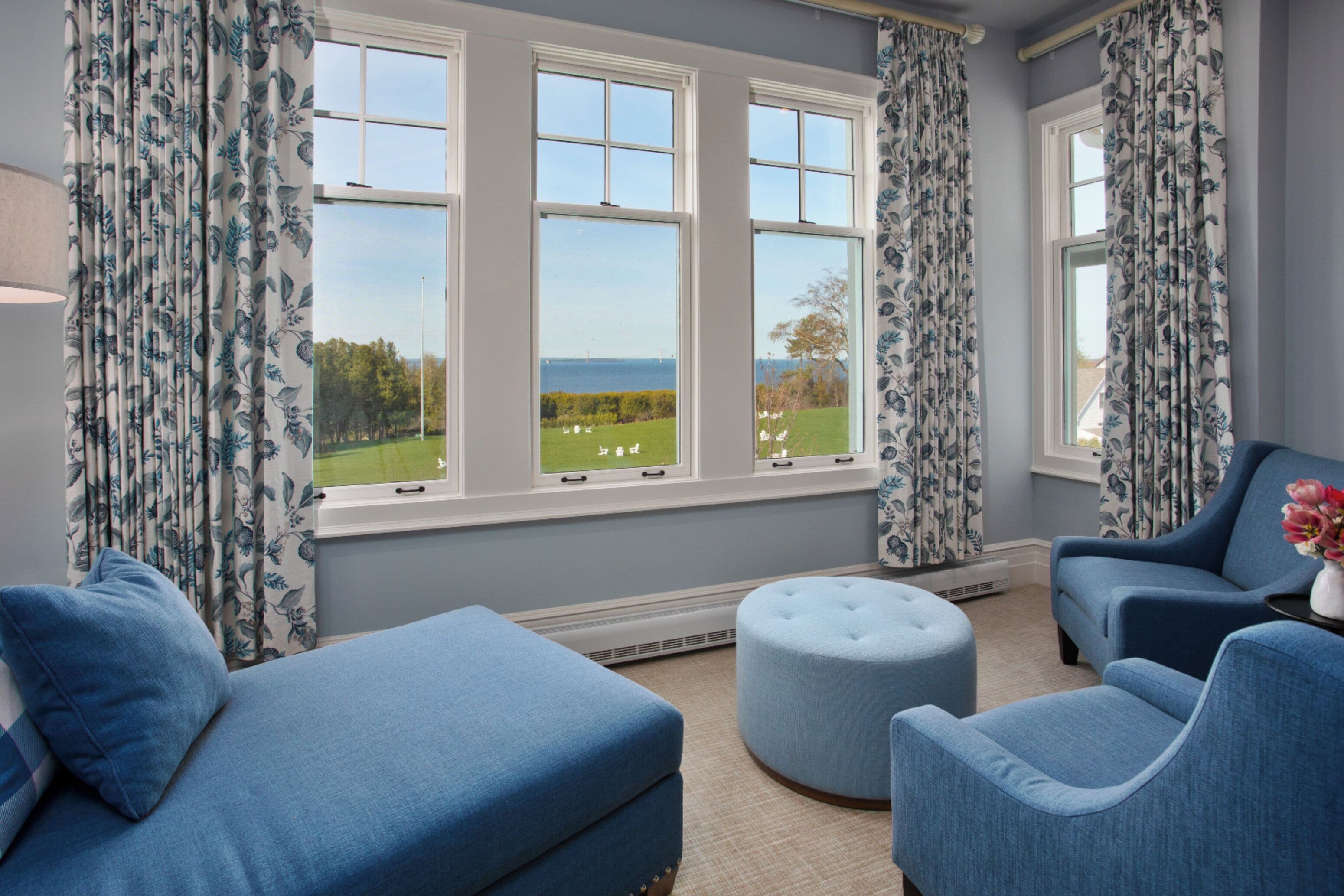 A room with a large window, a blue sofa, a round ottoman, and two blue armchairs.
