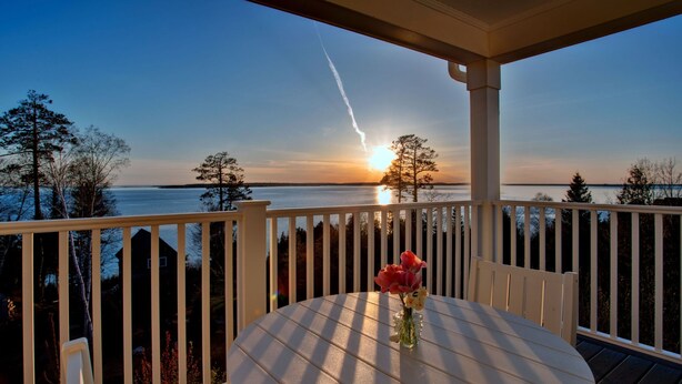 A deck with a round table and a vase of flowers, overlooking a body of water with trees and a sunset. - The Inn At Stonecliffe (Mackinac Island)