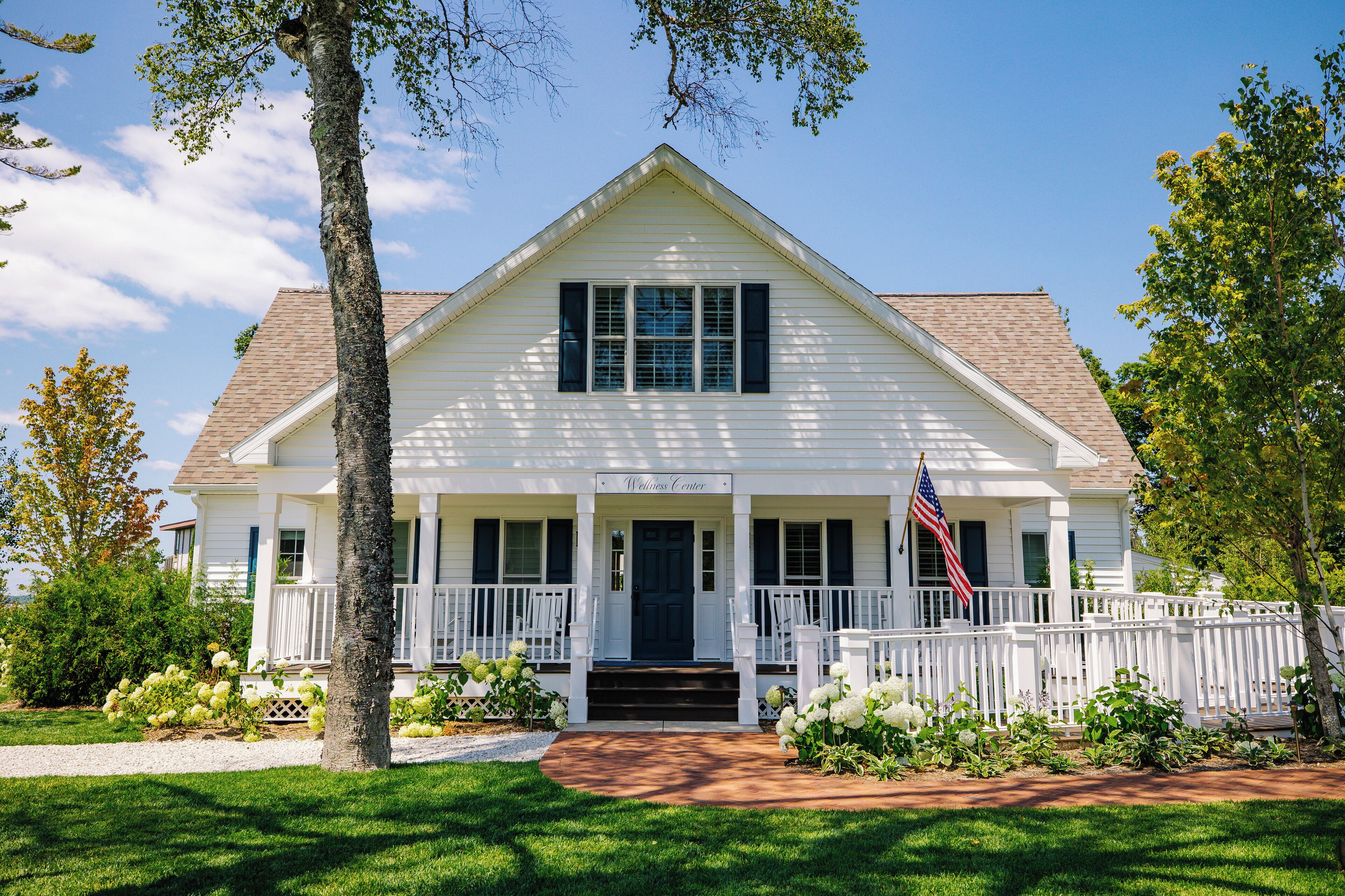 A white two-story house with a front porch, a small garden with white flowers, and an American flag.