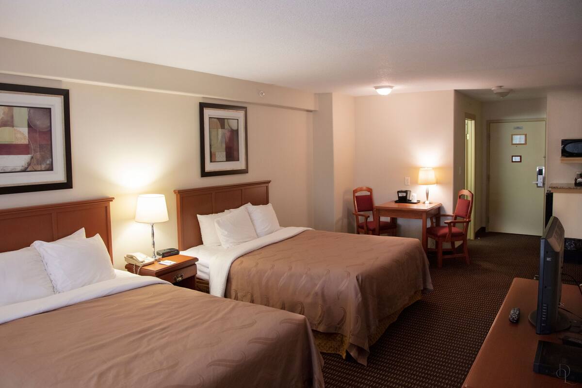 Quarto de hotel com duas camas, uma escrivaninha com luminária, uma cadeira, televisão e quadros nas paredes.