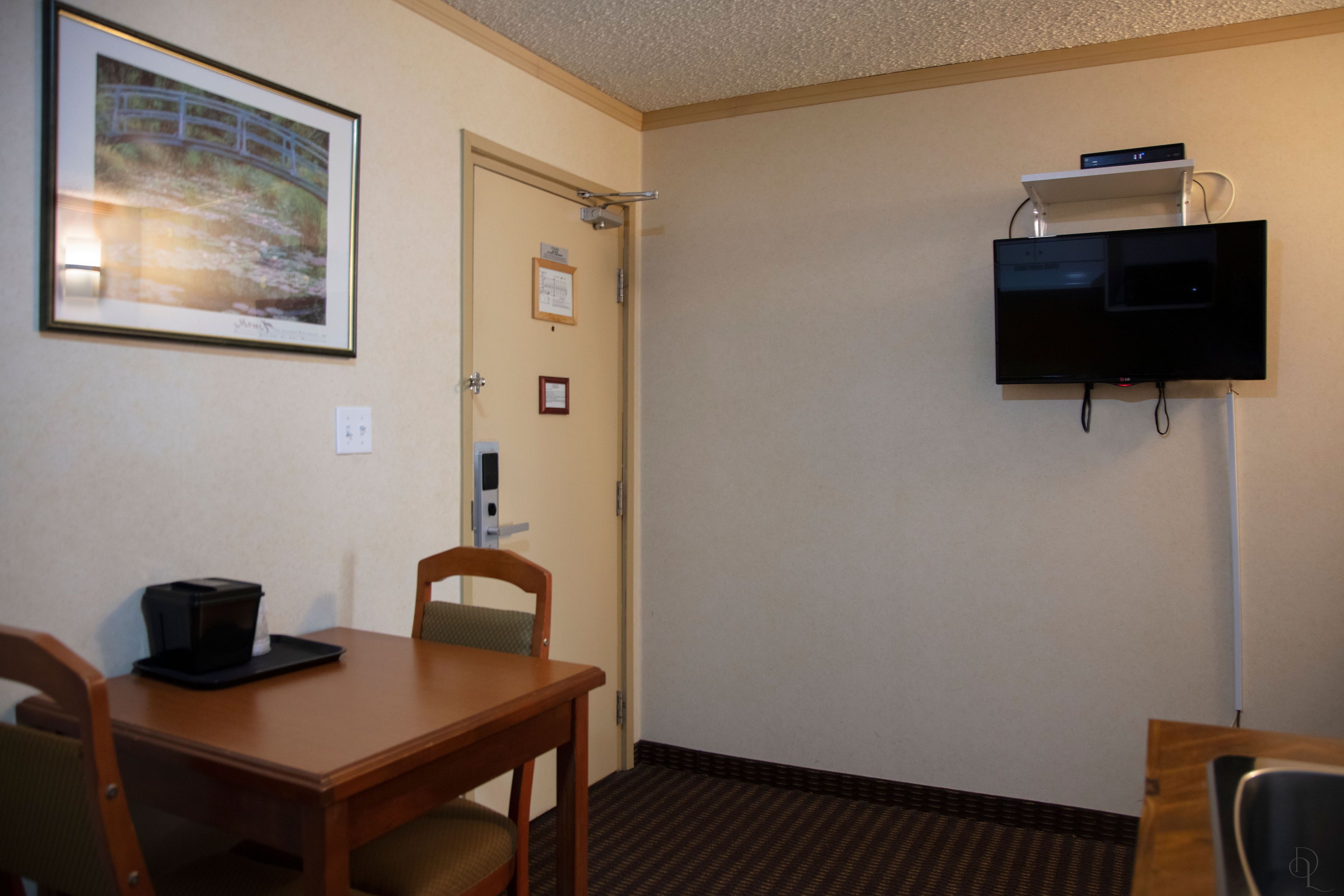 A hotel room with a wooden table, chairs, a framed picture on the wall, a television mounted on the wall, and a door with a sign.