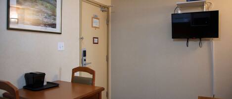 A hotel room with a wooden table, chairs, a framed picture on the wall, a television mounted on the wall, and a door with a sign.