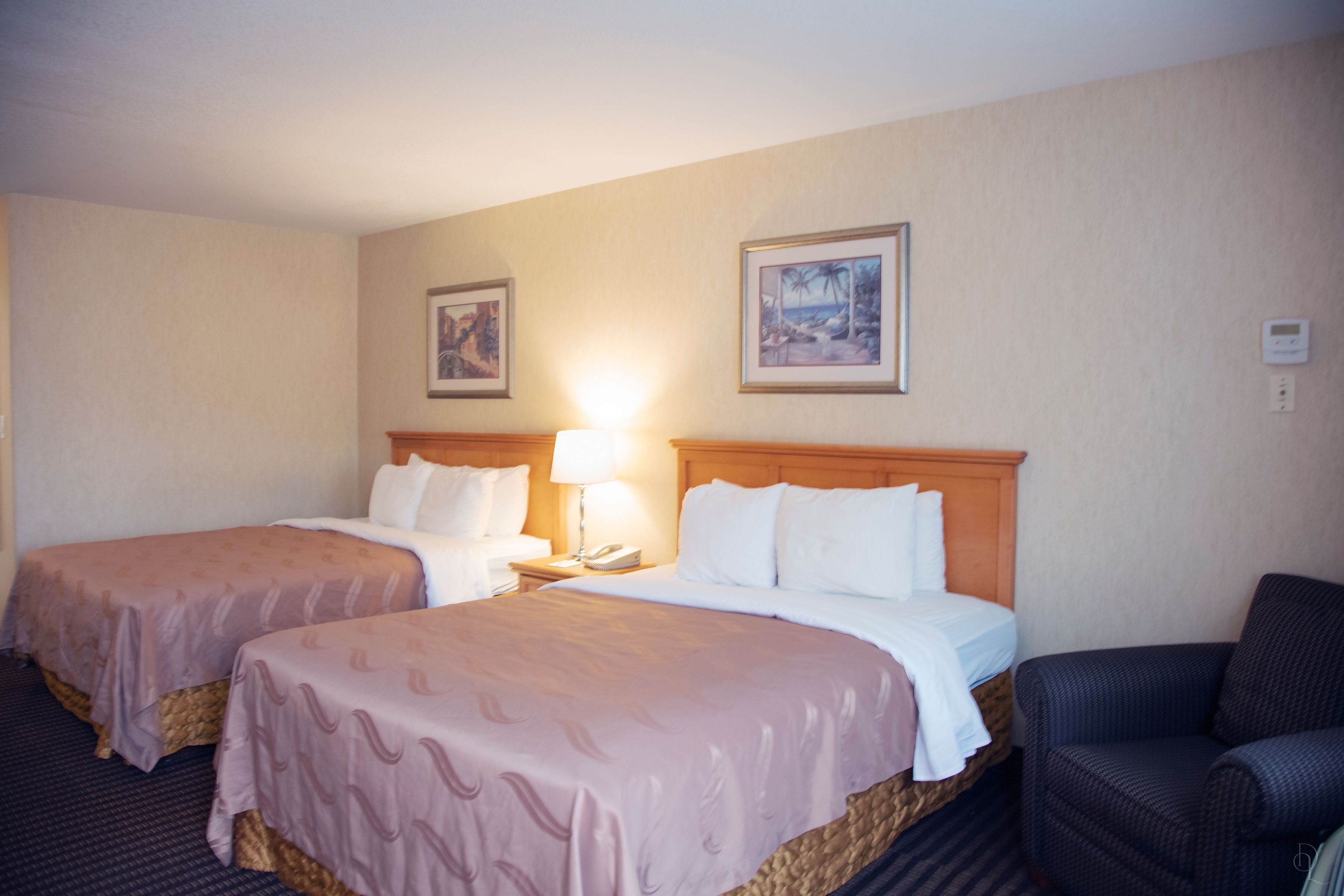 A hotel room with two beds, a nightstand with a lamp, a chair, and two framed pictures on the wall.