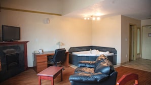 A room with a leather sofa, a desk with a computer, a television, a fireplace, and a large bathtub. - All Stay Suites (Westlock)