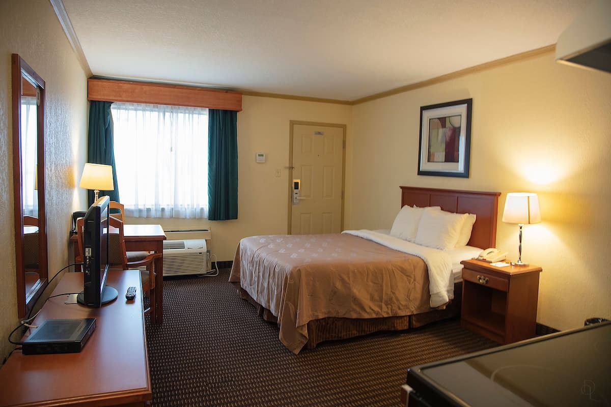 Quarto de hotel com cama, mesa de cabeceira com abajur, escrivaninha com computador e telefone, uma cadeira e janela com cortinas.