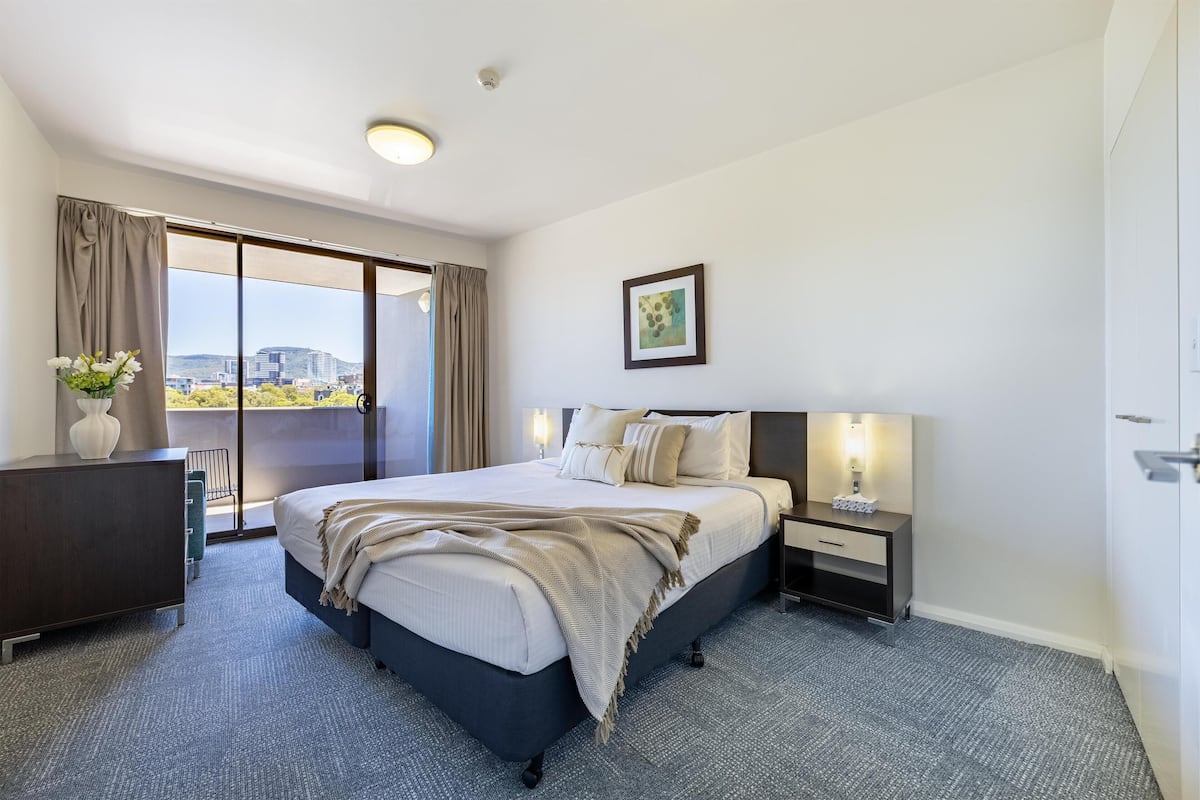 A hotel room with a large bed, a desk, a chair, a view of the city, and a balcony.