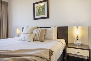 A neatly made bed with a beige blanket and pillows, a bedside table with a lamp, and a framed artwork on the wall.
