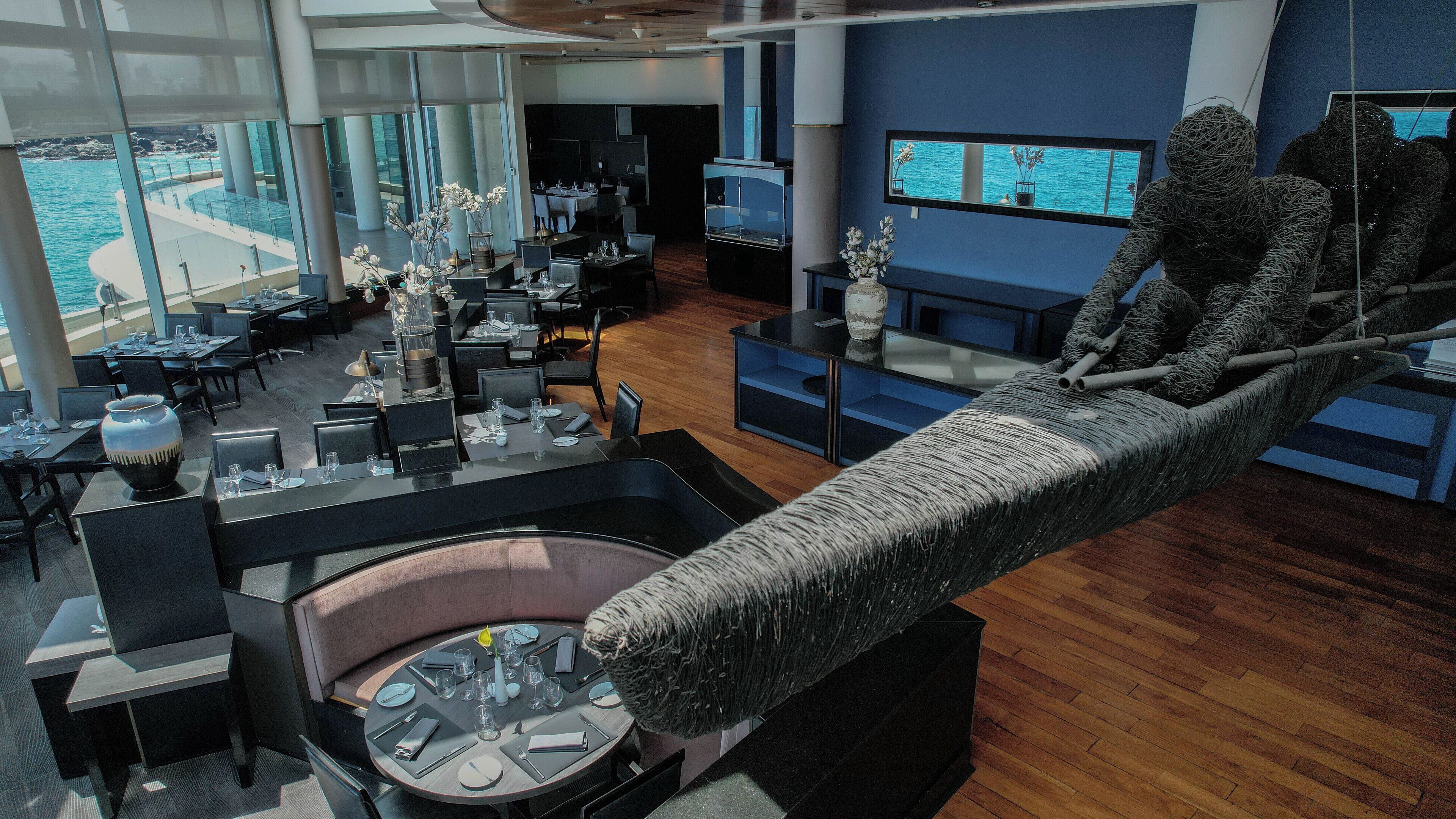 A modern restaurant with a view of the ocean, featuring round tables, black chairs, and a unique hanging sculpture.