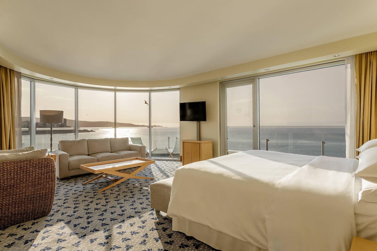 Habitación de hotel con cama, sofá, una mesita y televisor. La habitación tiene ventanas amplias con vista al mar.