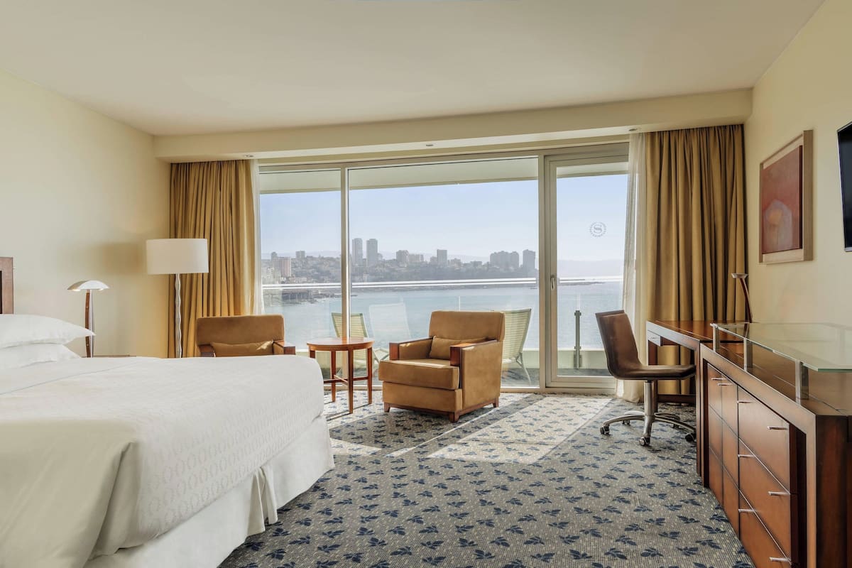 Habitación de hotel con cama, dos sillones, un escritorio y un ventanal con vista a la ciudad.