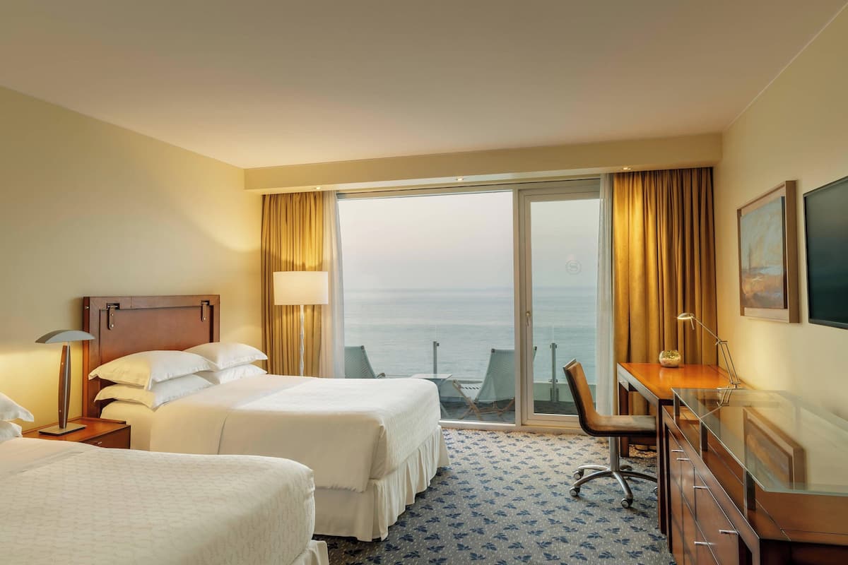 Habitación de hotel con dos camas, un escritorio, una silla, un televisor y vista al mar a través de una puerta corrediza.