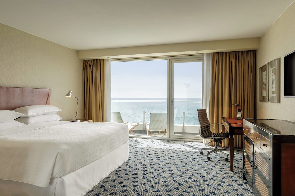 Habitación de hotel con una cama grande, un escritorio, una silla y vista al mar a través de una puerta corrediza de vidrio.