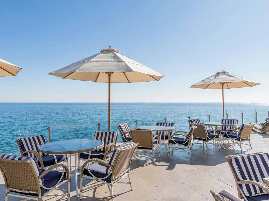 Een terras met rieten stoelen en tafels, grote parasols en uitzicht op zee.